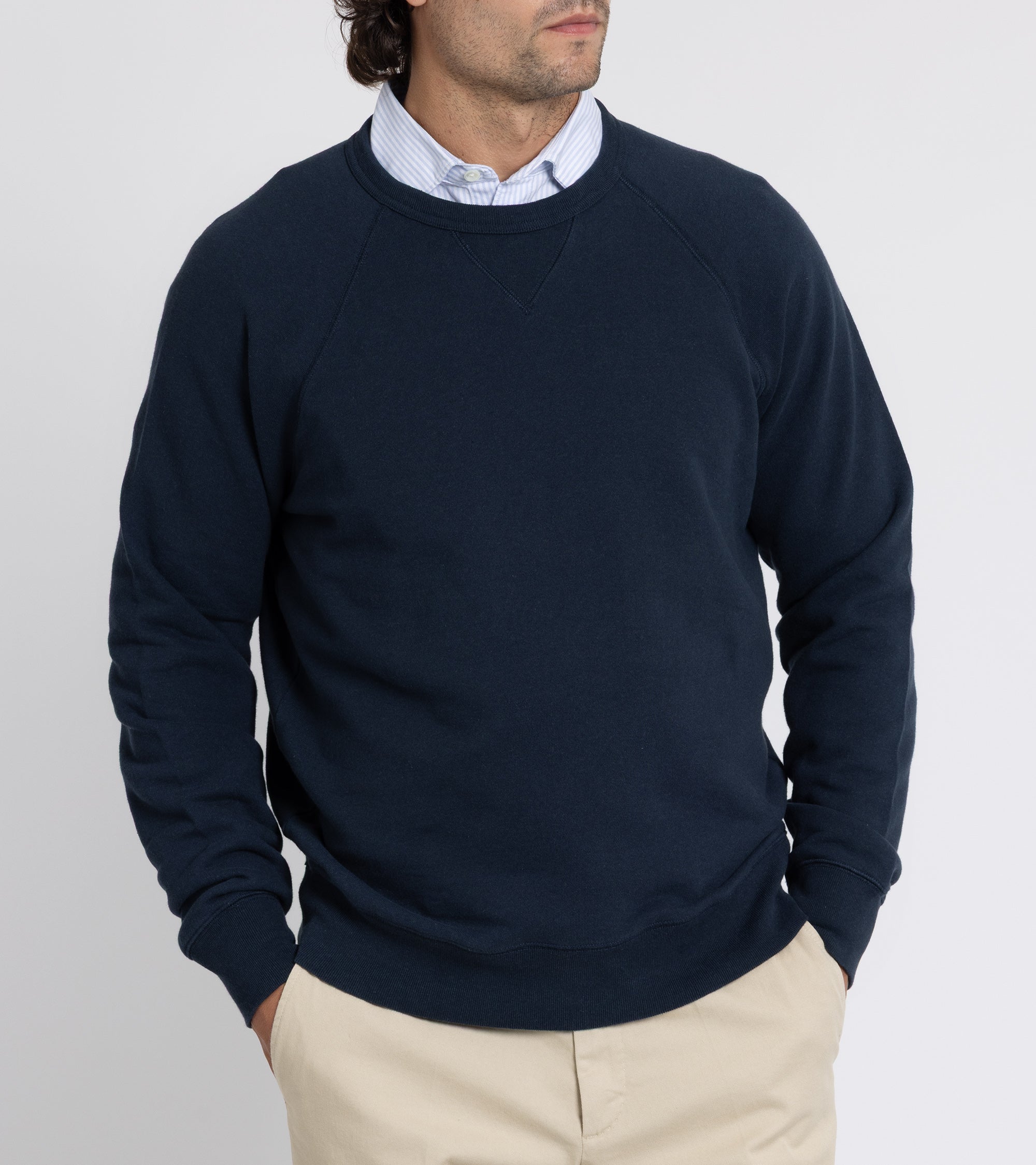 Man wearing a navy sweater over a collared shirt with beige pants on a plain background